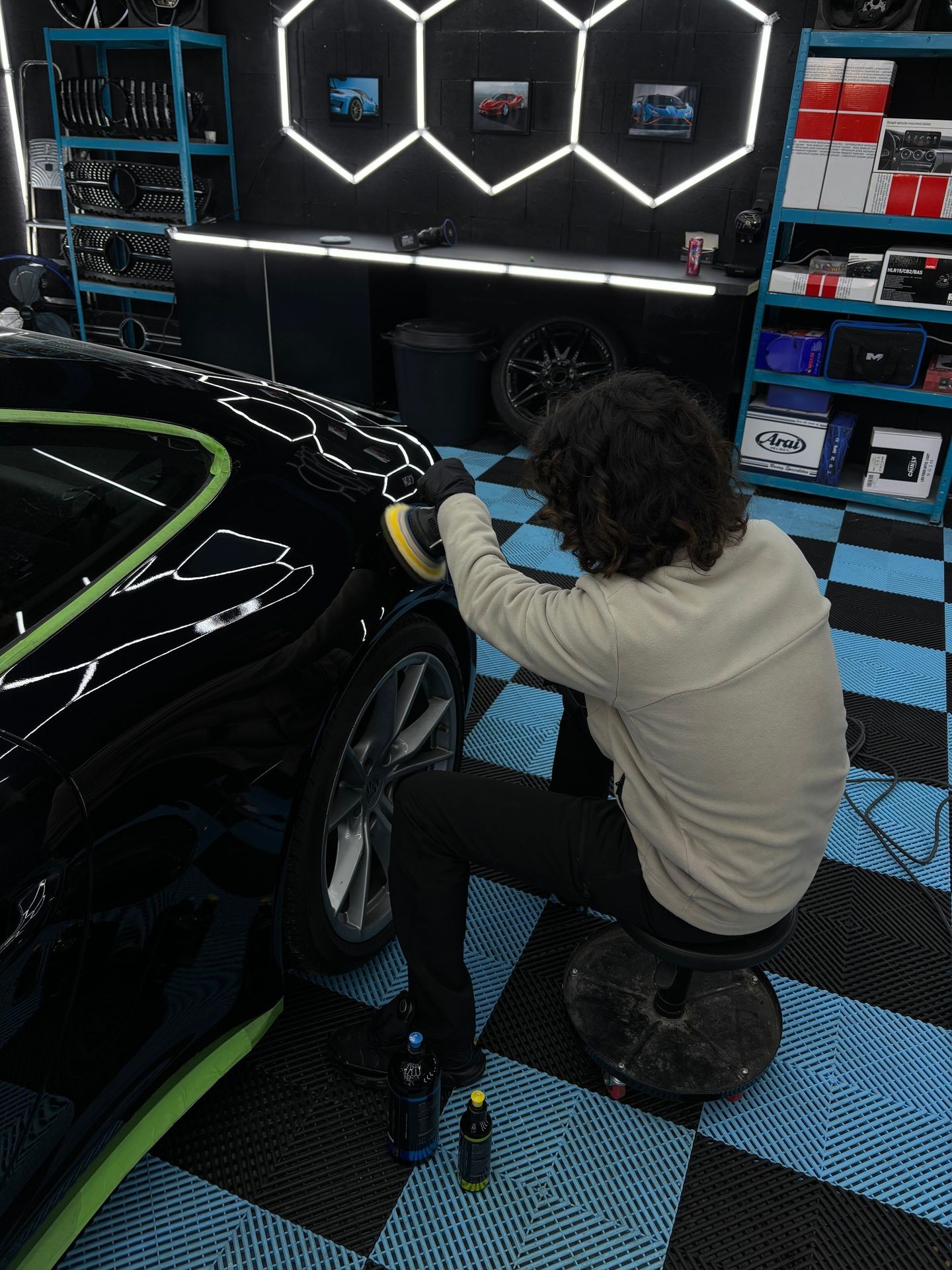 Person polishing a black car in a garage with geometric lights on wall, surrounded by automotive equipment.